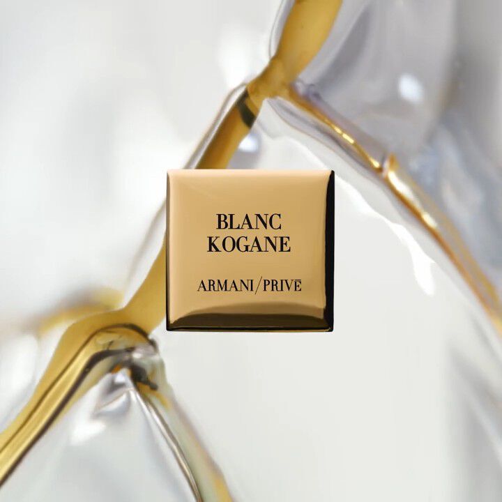 A close-up of a gold square perfume bottle with the label "BLANC KOGANE" and "ARMANI/PRIVE" prominently displayed. The background features a blurred, translucent material.