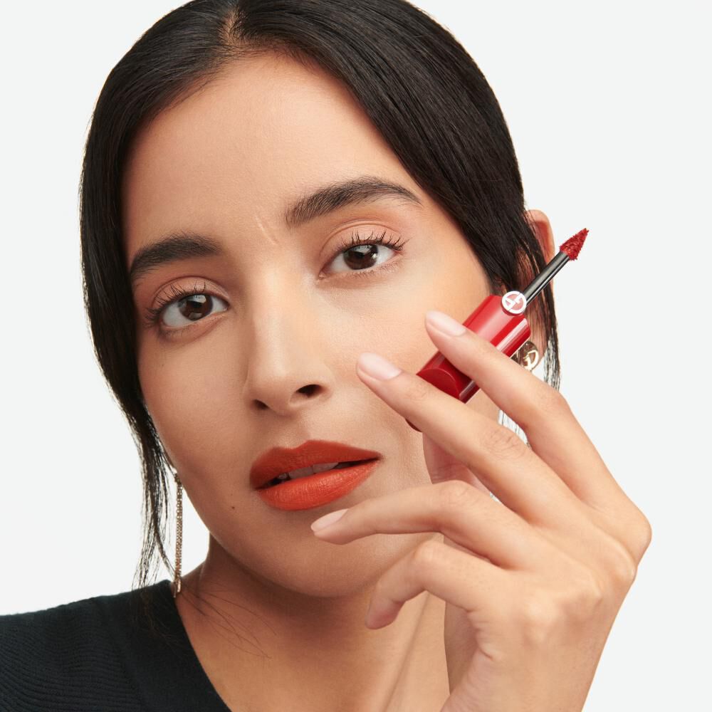 A person with dark hair and a subtle makeup look holds a red lipstick in one hand, looking directly at the camera with a confident expression.