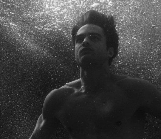 A black and white image of a man underwater, facing upwards with a focused expression, bubbles surrounding him.