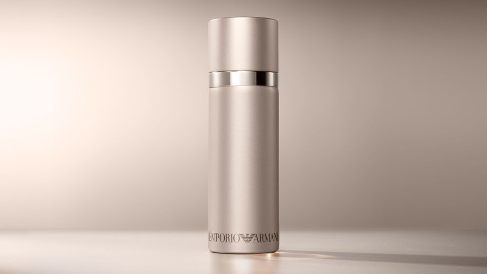 A matte rose cylindrical Emporio Armani "She" fragrance bottle is elegantly positioned on a warm, light brown background.