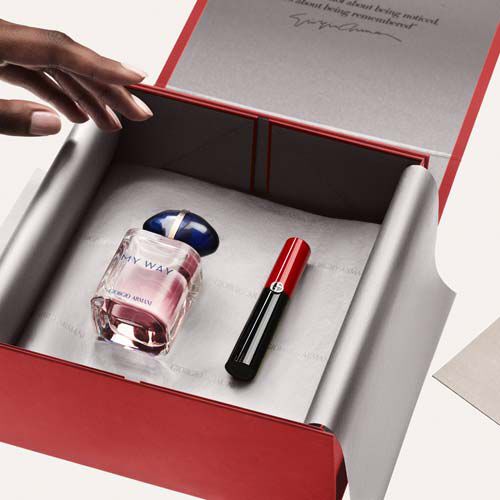 A hand reaches into an open red gift box containing a bottle of perfume and a lipstick. The box is lined with soft tissue paper.