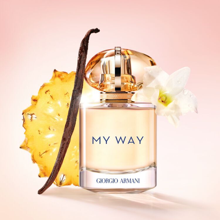 Armani My Way Sunny Vanilla perfume bottle displayed with a slice of pineapple, a vanilla bean, and tuberose flower, set against a soft pink background to highlight its fresh and natural ingredients.