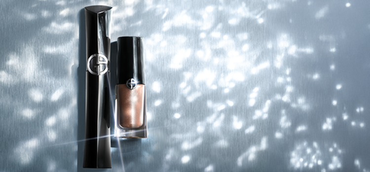 A woman with dark hair wears a bold purple eyeshadow and large black earrings, posing in a pink top. Beside her, two lip glosses in dark red and nude shades are displayed against a backdrop of elegant swirling colors.