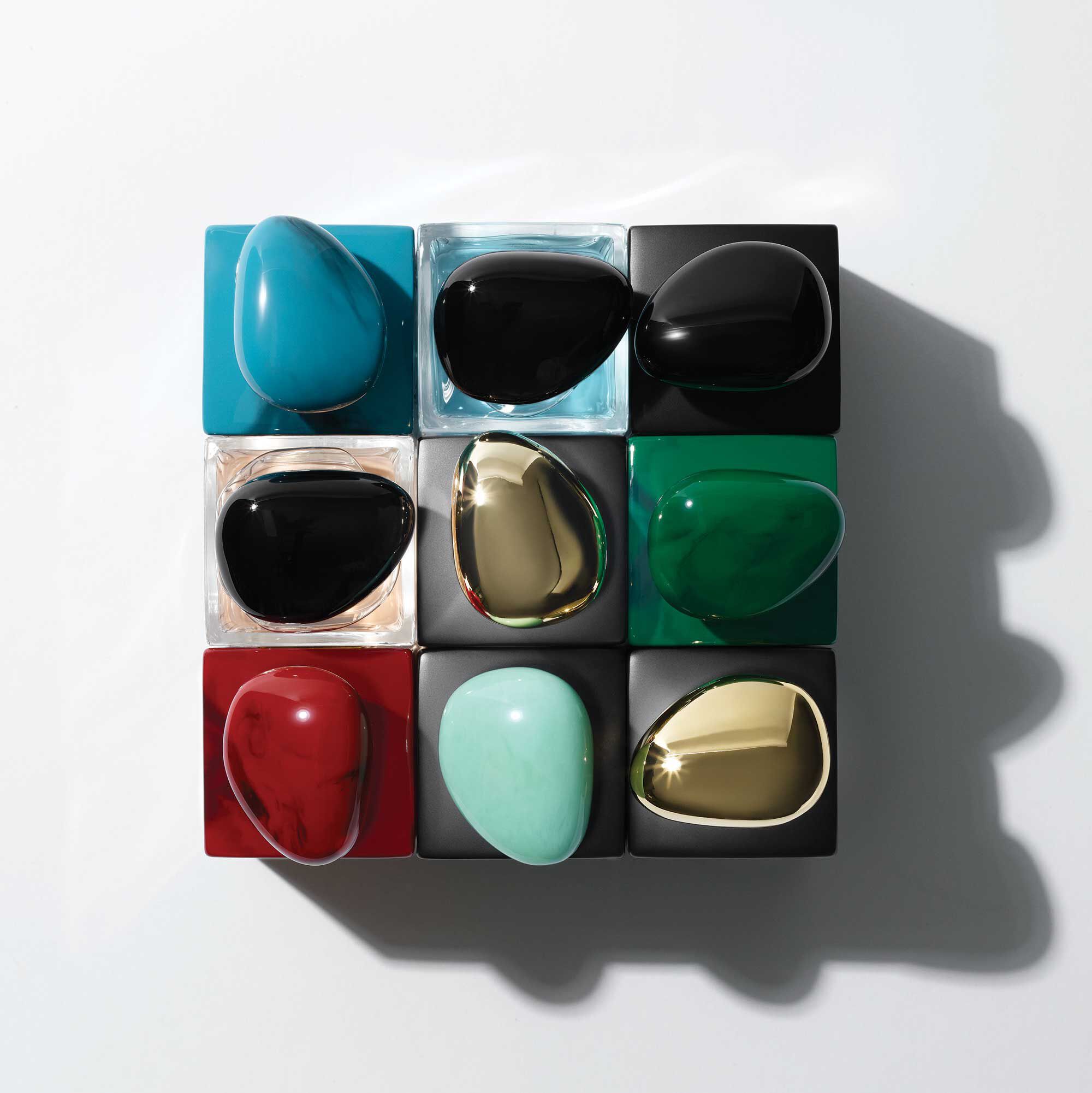 An arrangement of nine glossy, teardrop-shaped objects in various colors—black, turquoise, green, red, gold, and clear—set on a grid of black and colored squares against a white background.