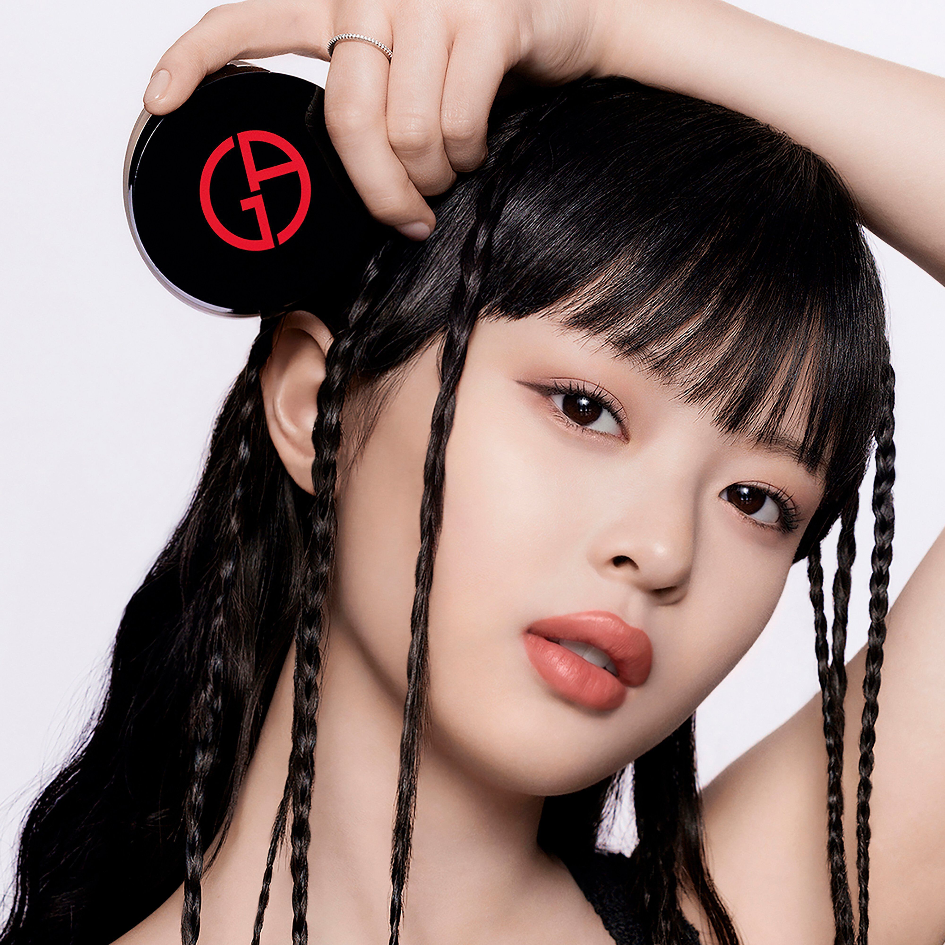 A young woman with black braided hair holds a round compact with a red logo, gazing confidently at the camera, wearing subtle makeup and a natural expression.