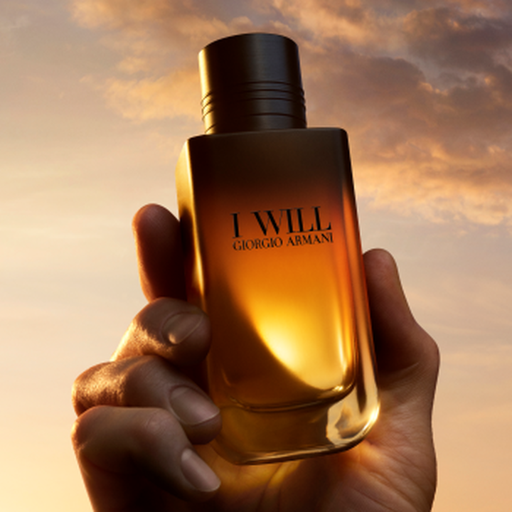Une main tenant le flacon de parfum I WILL Giorgio Armani devant un ciel dore au coucher du soleil.