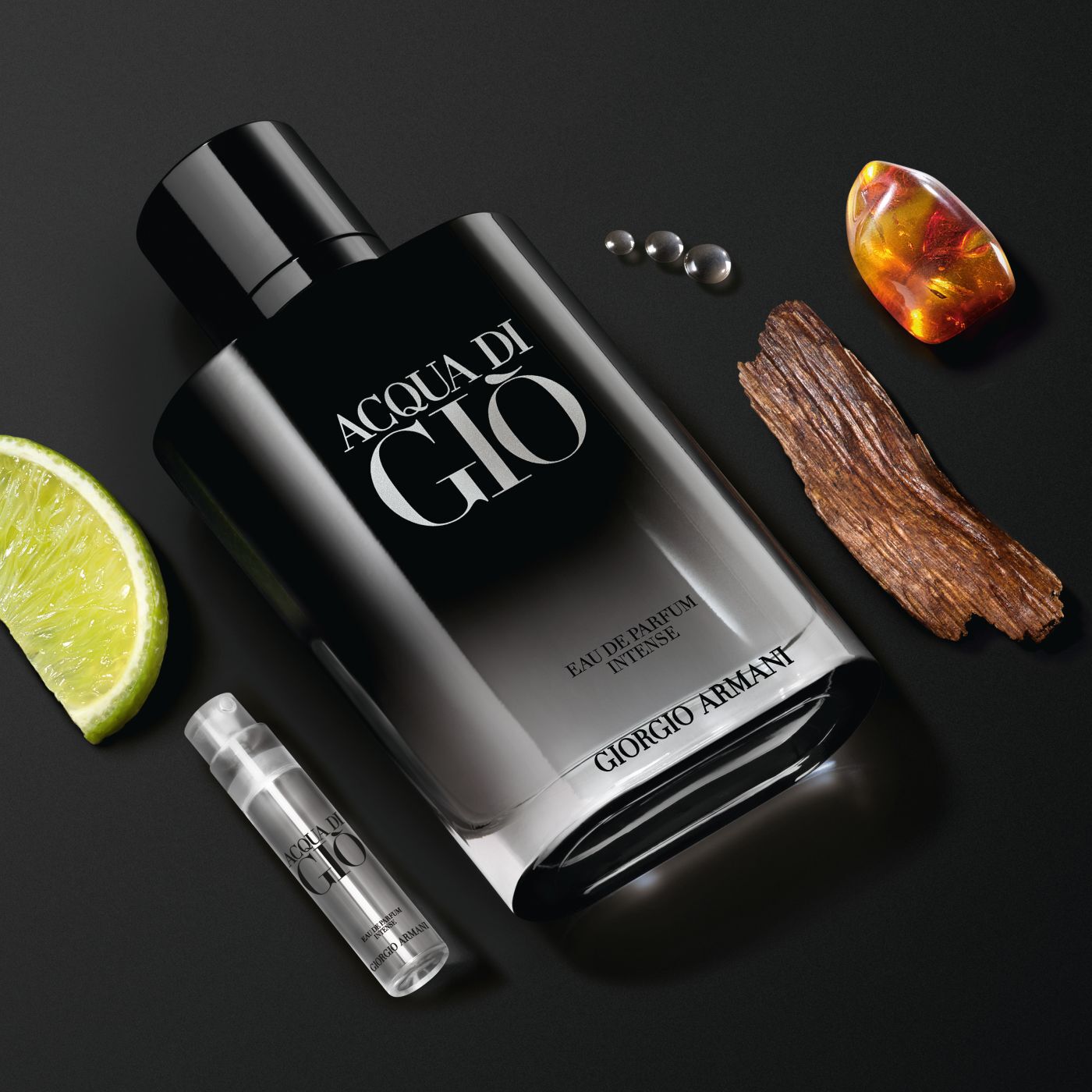 Acqua di Gio Eau de Parfum Intense Flakon mit Limette, Holz, Bernstein und Probeflaschchen auf schwarzem Grund.