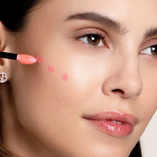 A close-up of a woman applying a coral pink blush to her cheek using a brush, with a natural makeup look and glossy lips.