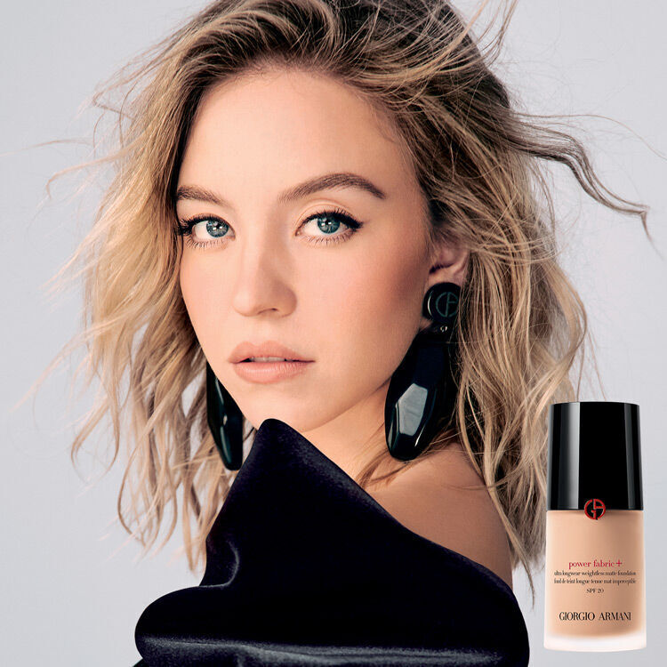 A striking model with wavy blonde hair and bold earrings poses on the left, while on the right is a bottle of Giorgio Armani foundation against a soft beige background.