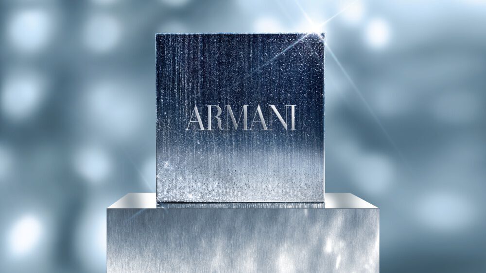 A square Armani GIFT SET in a shimmering silver and blue gradient box, displayed on a reflective pedestal with a soft, sparkling light background.