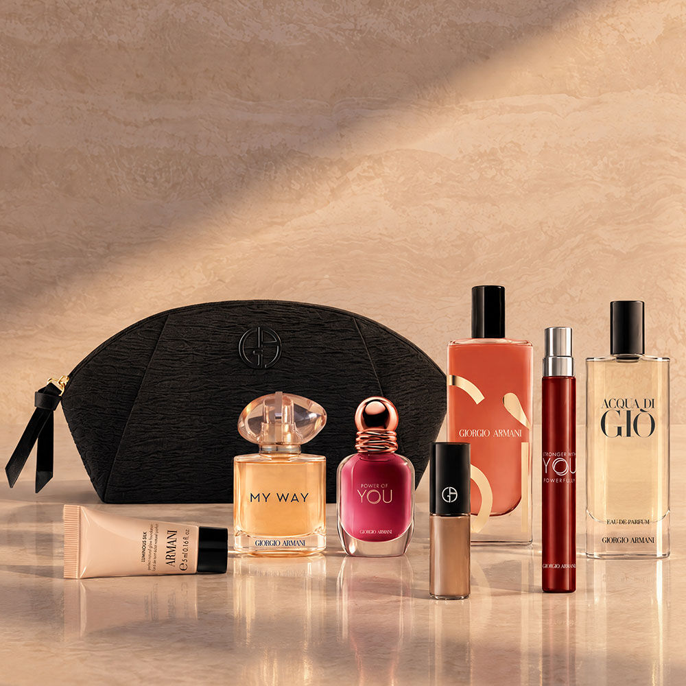 An Armani summer gift set presented on a white marble ledge overlooking a calm blue sea. At the center is a beige knit pouch featuring a blue embroidered GA logo. On the left are Si Passione, Acqua di Gio, a Luminous Silk foundation sample, a liquid blush, and a nude lipstick. On the right are Armani Code, My Way, and Power of You fragrance miniatures.