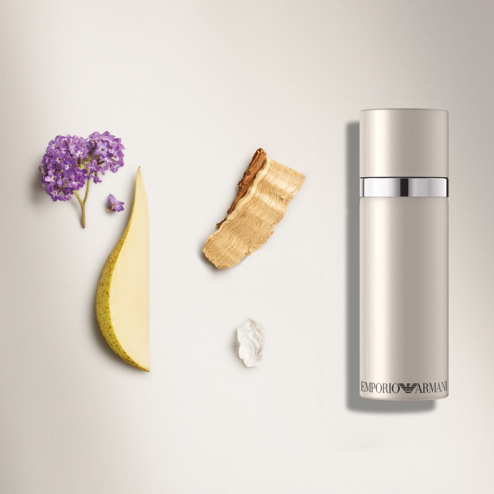 A close-up flat lay composition for Emporio Armani "She" fragrance, showing a light-beige bottle surrounded by ingredients like heliotrope accord (purple flowers), cedar wood, a sliced pear, and musk.