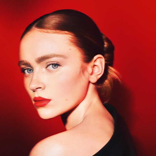 Portrait d'une femme aux cheveux blonds, avec un fond rouge eclatant, mettant en valeur son maquillage elegant et son regard captivant.