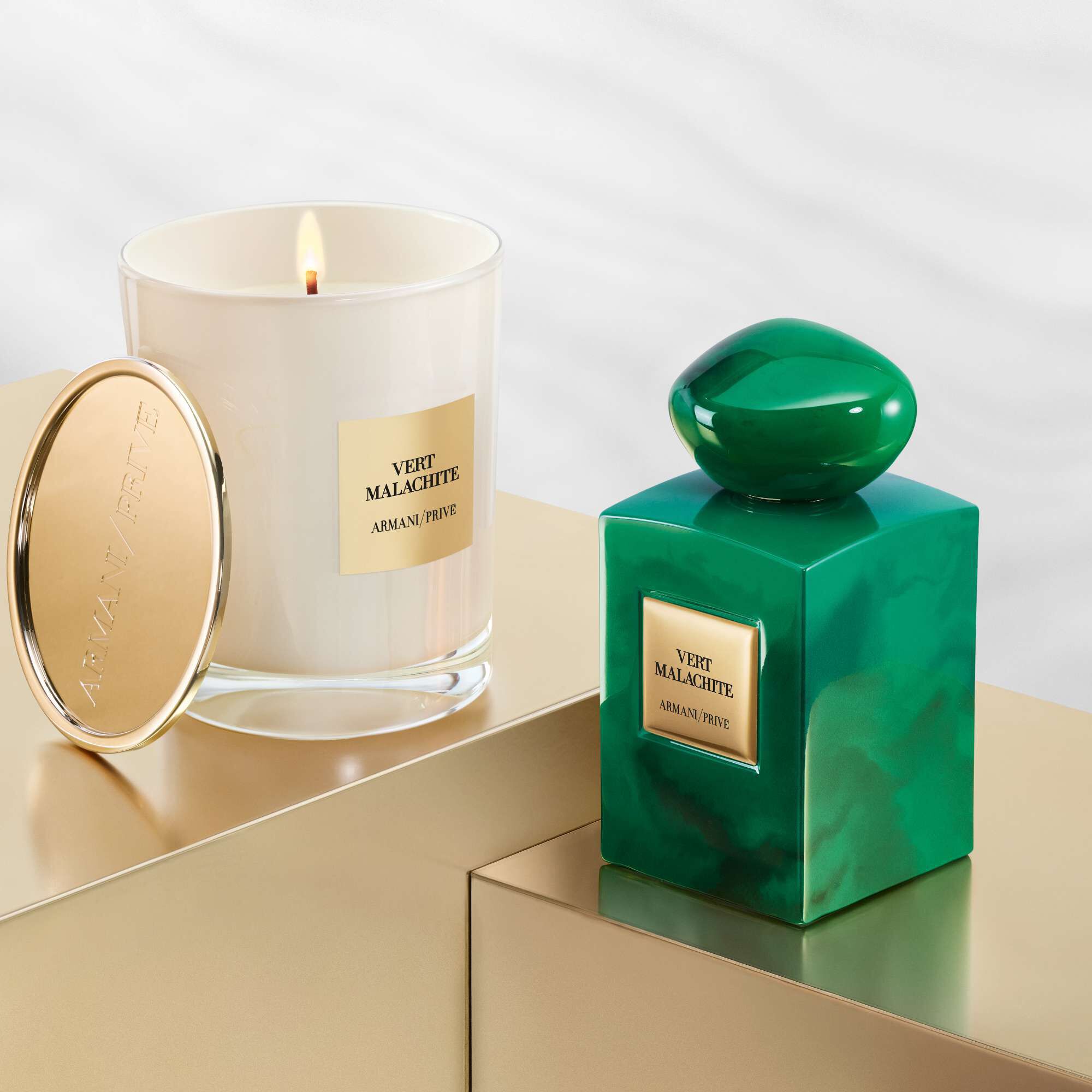 A luxurious green candle next to a matching green perfume bottle, both elegantly displayed on a gold surface, featuring a label that reads "Vert Malachite" by Armani Prive.