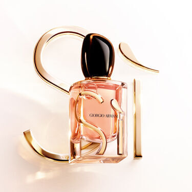 A smiling woman with short blonde hair is wearing a black off-shoulder dress, standing against a vibrant red background. To the right, a bottle of perfume is displayed beside the text "Si" in a stylish font.