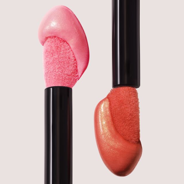 Two applicators with glossy pink and coral lip products, set against a soft neutral background.