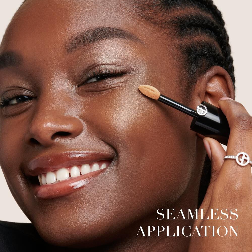 A smiling person with braided hair applies foundation using a small sponge applicator, showcasing a flawless skin tone. The text "SEAMLESS APPLICATION" is featured at the bottom.