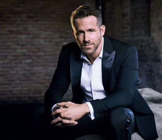 A man in a tuxedo is seated, looking confidently at the camera with a slight smile. The background features brick walls, and the lighting is soft, giving a warm ambiance to the portrait.