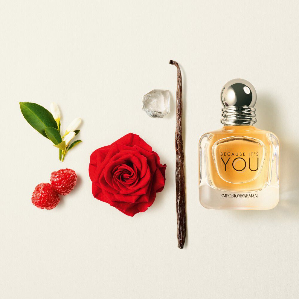 The clear glass bottle of Emporio Armani "Because It's You" fragrance is featured with a red rose, vanilla bean, raspberries, a white flower, and an ice cube on a white background.