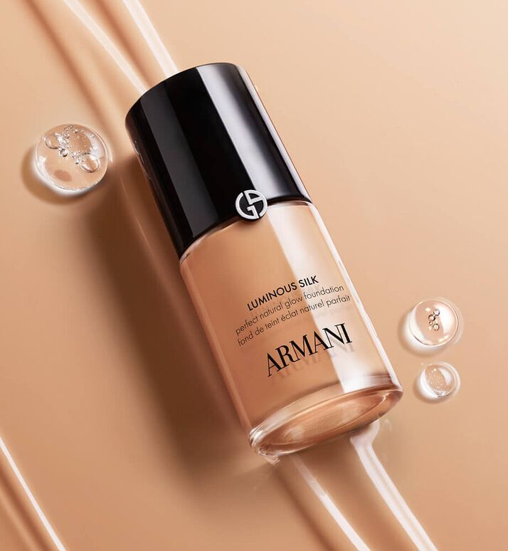 Bild einer Armani Luminous Silk Foundation-Flasche in beiger Verpackung, umgeben von Tautropfen auf golden-beigem Hintergrund.