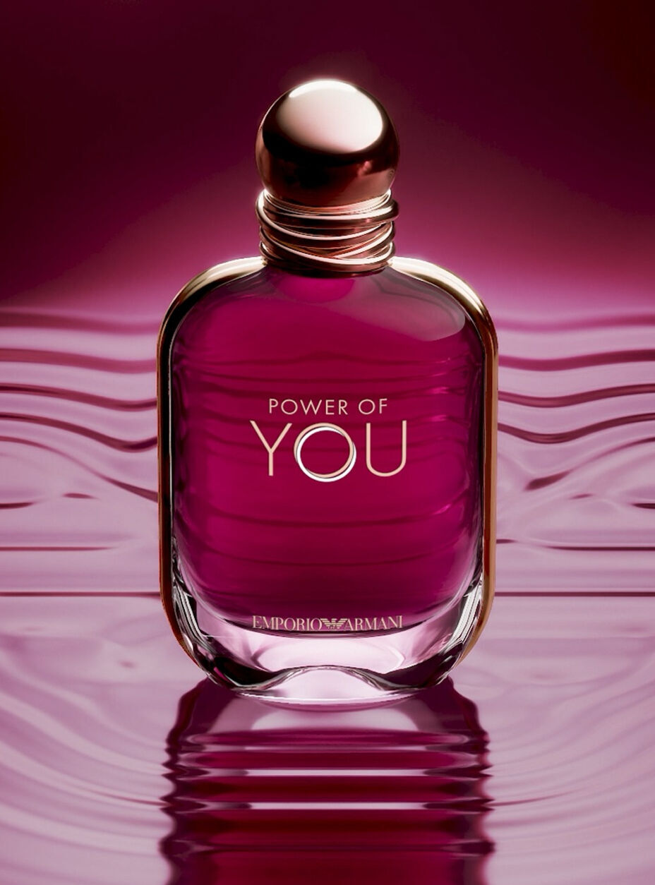 Une publicite en gros plan pour le flacon de parfum « Power of You » d'Emporio Armani. Le flacon en verre aux teintes rubis est place au centre d'une surface liquide magenta fonce reflechissante qui presente de legeres ondulations. Le flacon possede un bouchon dore arrondi et les inscriptions « POWER OF YOU » et « EMPORIO ARMANI » imprimees en lettres dorees. L'arriere-plan est un degrade doux et flou de rose profond et de violet. En bas, un texte blanc indique : « Presentation d'un nouveau flacon emblematique : un bijou aux tons rubis », suivi d'un lien « DECOUVRIR » et d'une icone de pause.