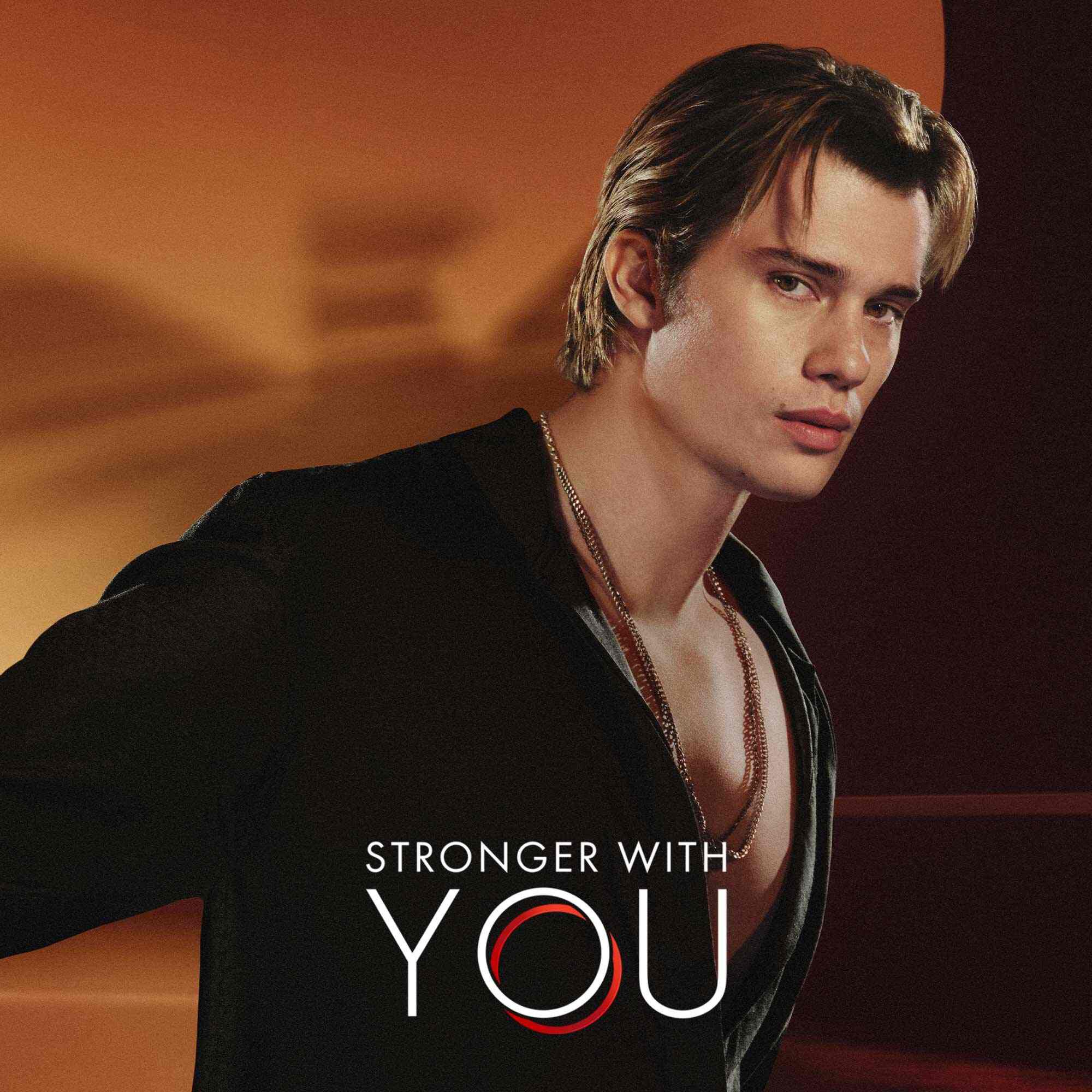 Nicholas Galatzine en chemise noire avec le texte du logo 'Stronger With You' sur fond orange chaud