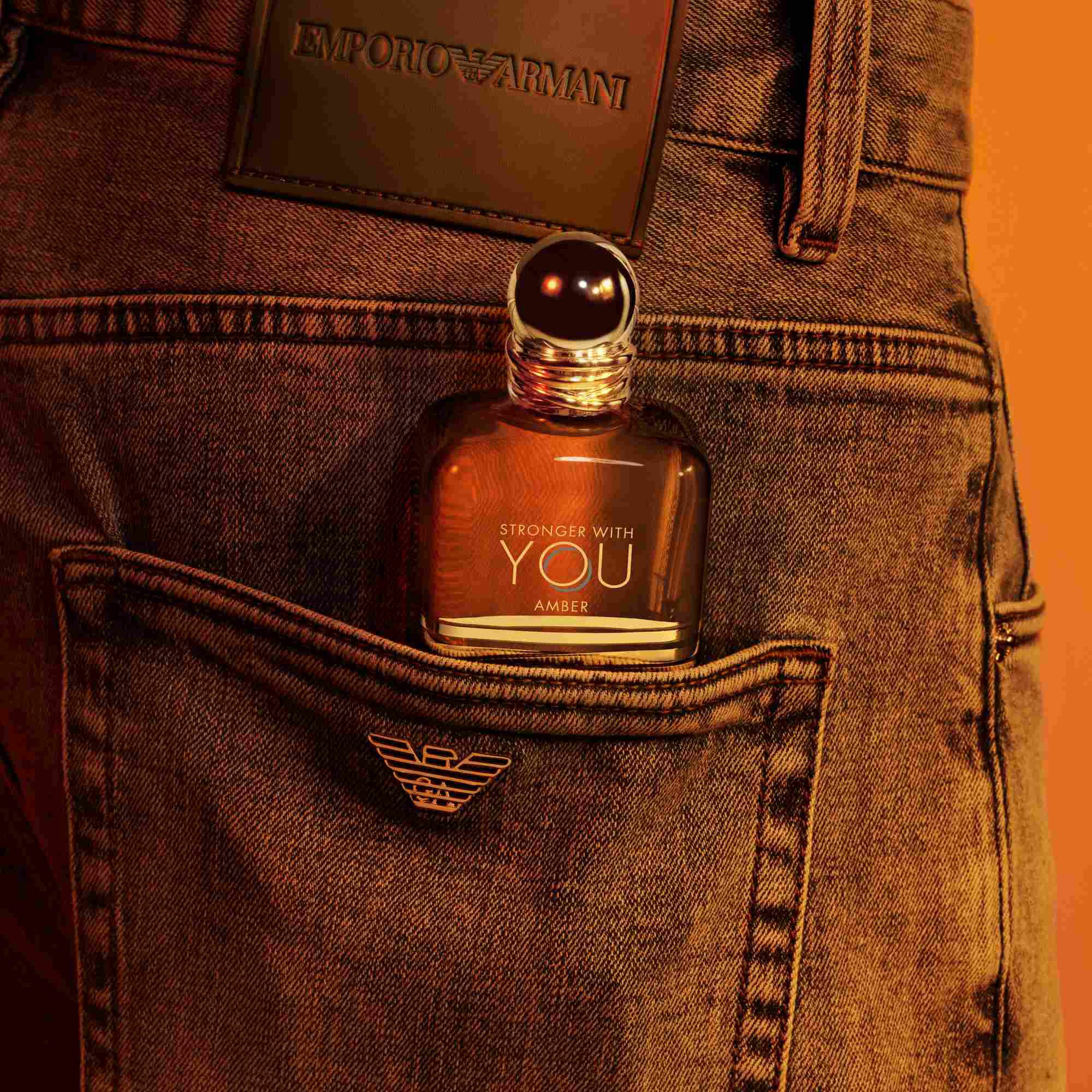Un flacon de parfum Emporio Armani Stronger With You Amber depassant de la poche arriere d'un jean, avec un patch en cuir fonce visible au-dessus