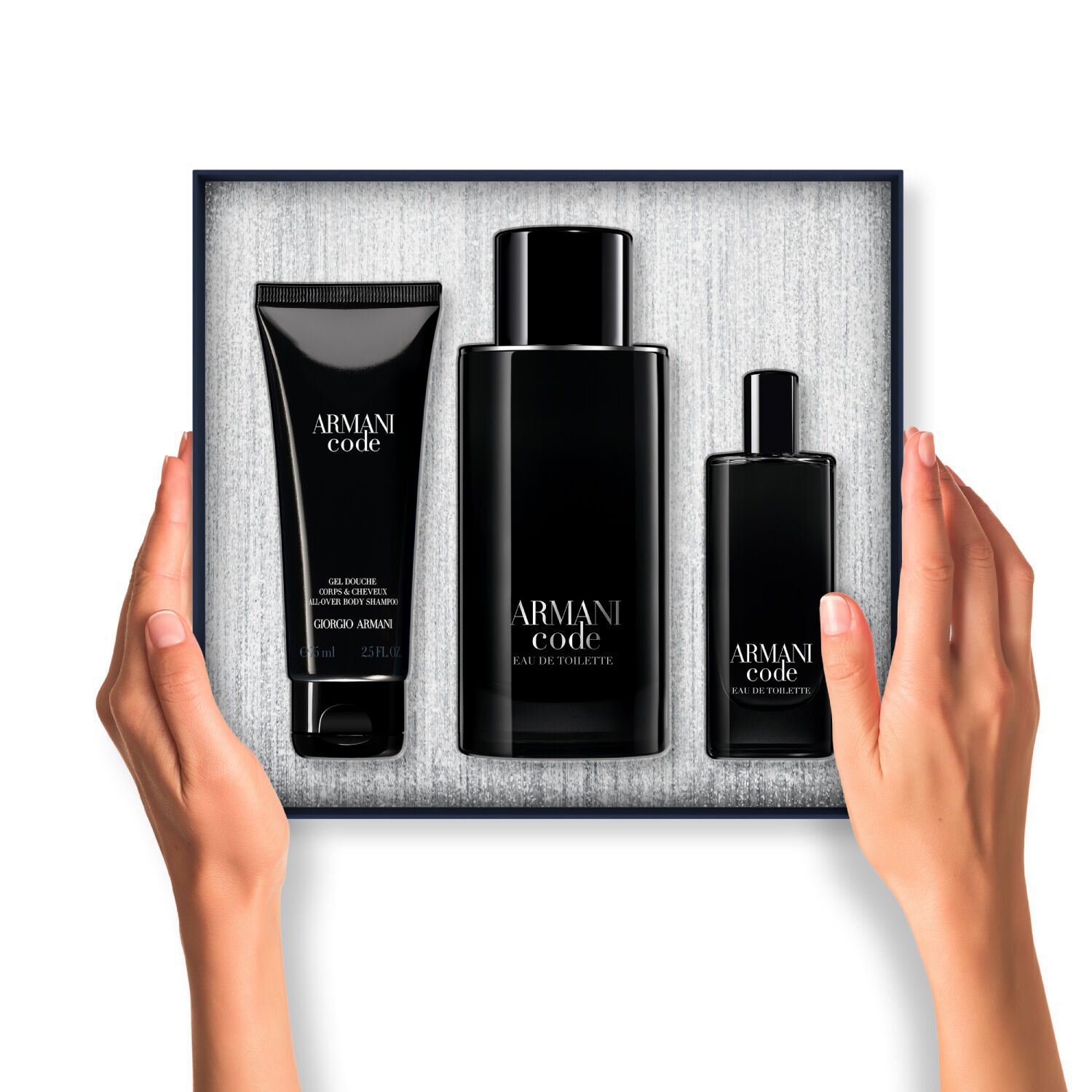 Une paire de mains tient un coffret cadeau Armani Code ouvert. A l'interieur, trois produits emballes en noir (gel douche, grande Eau de Toilette et Eau de Toilette format voyage) sont soigneusement disposes sur un fond argente texture.
