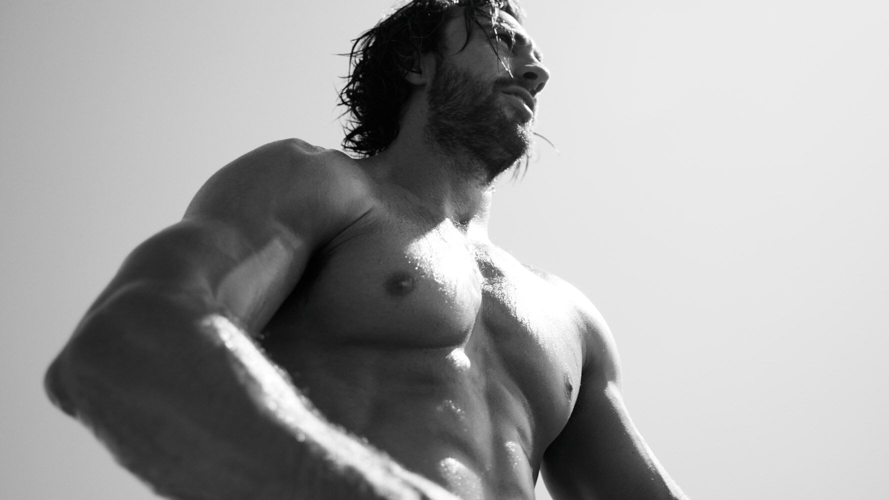 Ein muskulöser Mann steht am Strand, mit nassem Oberkörper und intensivem Blick, im monochromen Schwarz-Weiß.