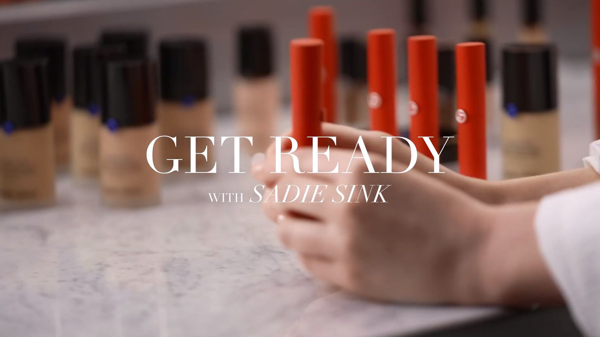 A close-up of hands preparing makeup products on a marble counter, featuring foundation bottles and the text 'Get Ready with Sadie Sink.'